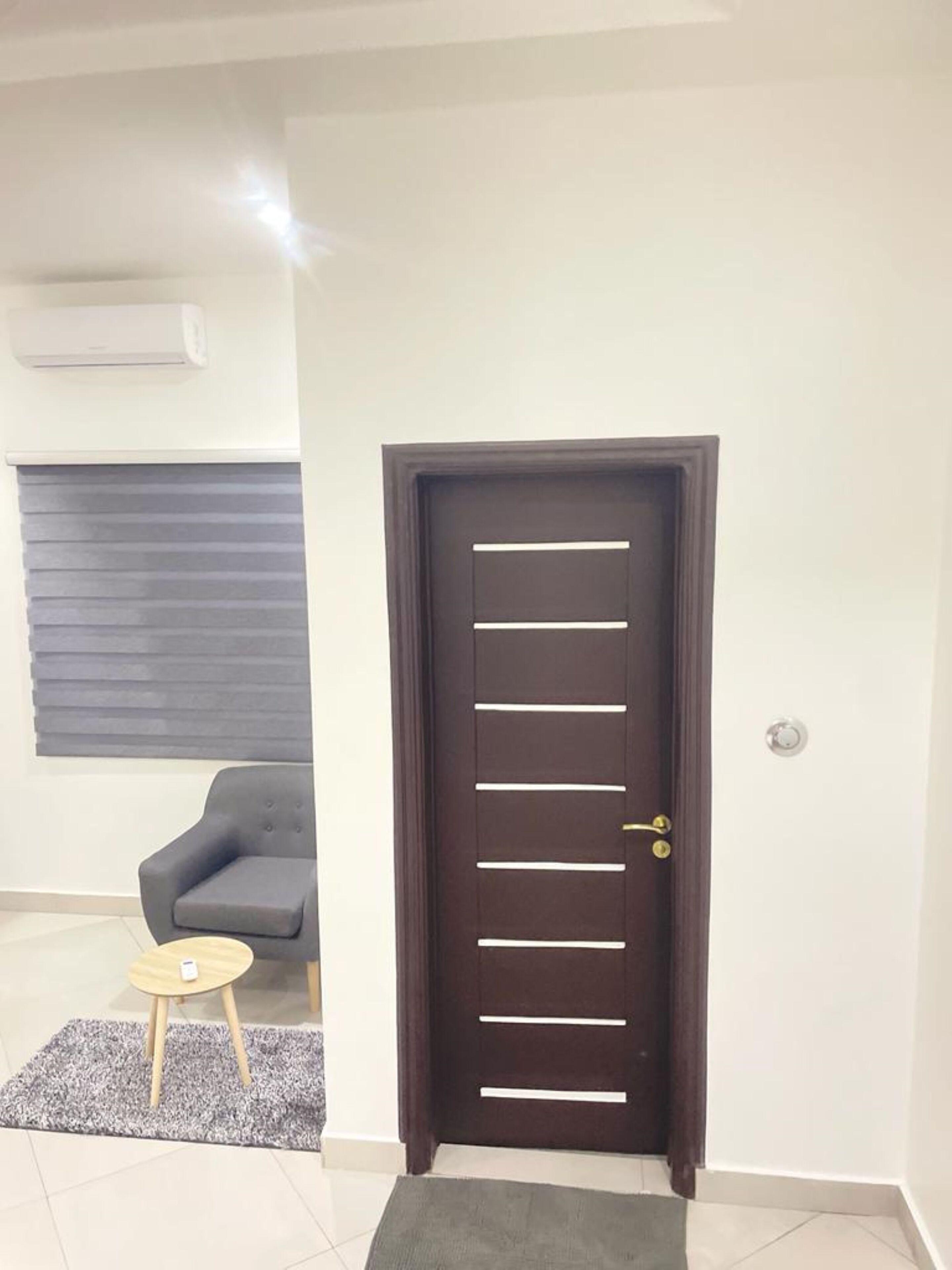 Una puerta de madera oscura con manijas horizontales, un tapete gris, una pequeña mesa redonda y un sofá gris en una habitación con paredes blancas y una lámpara en el techo.