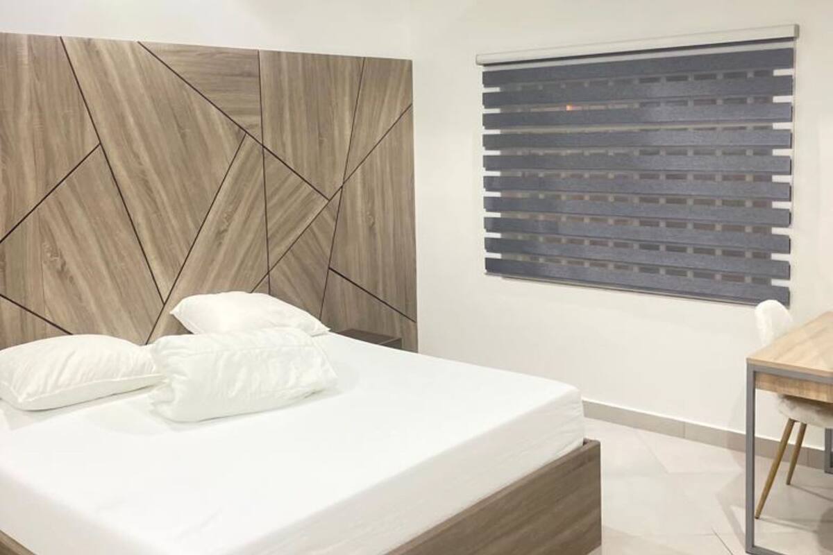 A modern bedroom with a wooden headboard, a bed with white bedding, a ceiling fan, and a window with blinds.