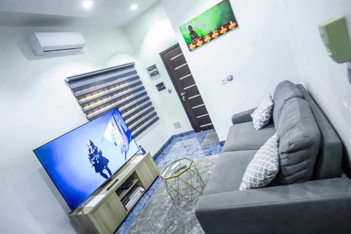 A living room with a flat-screen TV, a grey sofa, a small coffee table, and a window with blinds.