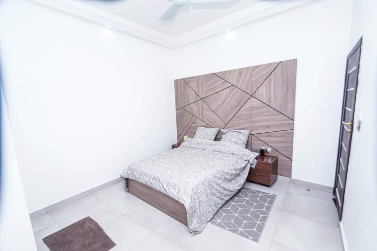 A modern bedroom with a geometric-patterned headboard, a neatly made bed, a nightstand, and a small wooden cabinet.