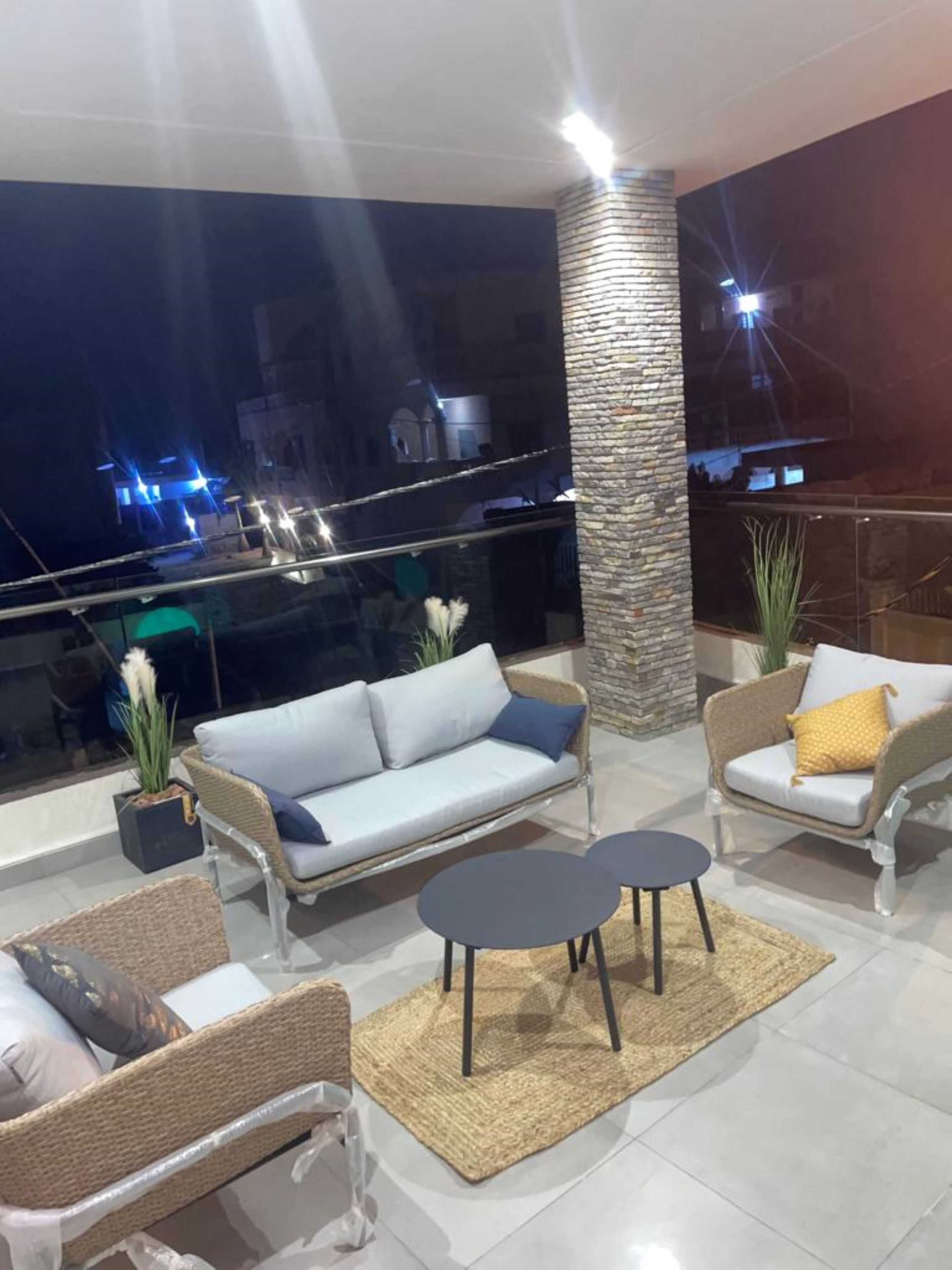 Un espacio exterior moderno con mobiliario de mimbre, un pilar de piedra y vista a la ciudad de noche.