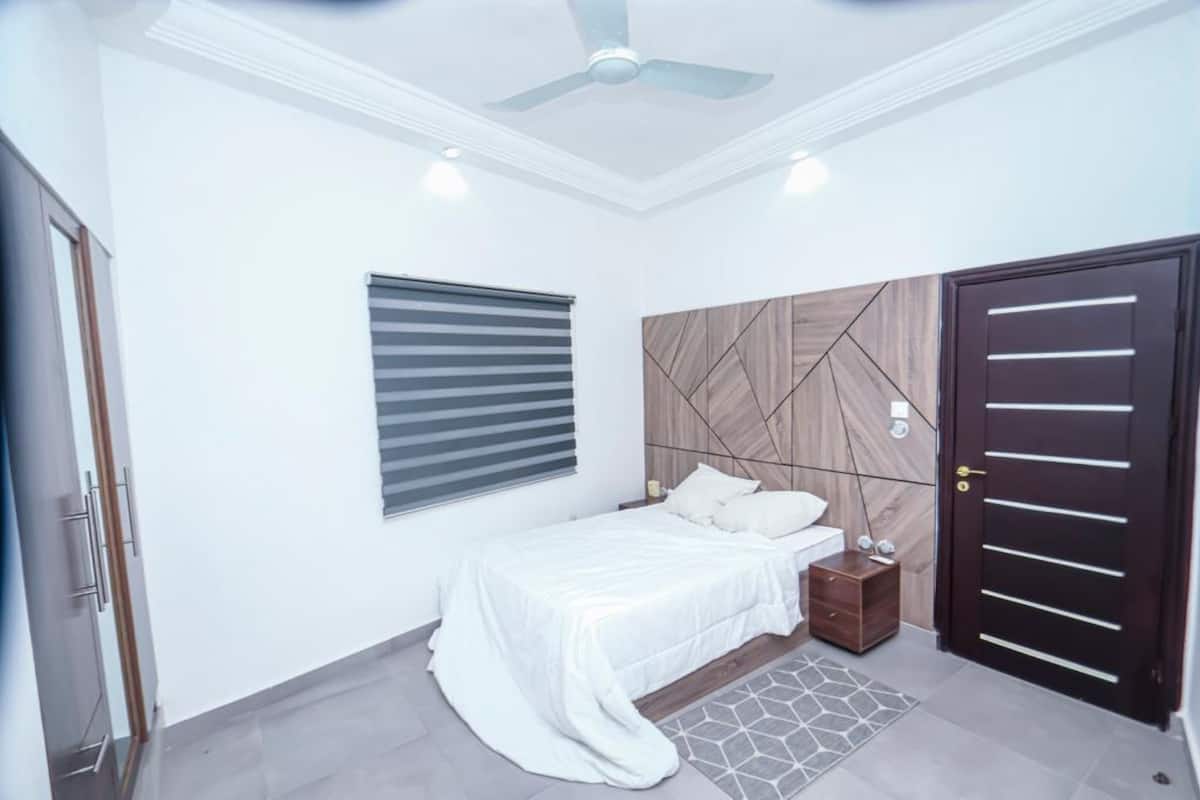 A bedroom with a bed, bedside tables, a window with blinds, a ceiling fan, and a door.