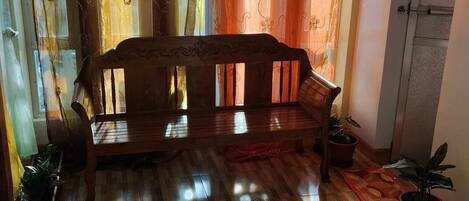 Un banc en bois dans une pièce dont le sol est recouvert de planches de bois, avec de grandes fenêtres ornées de rideaux et des plantes en pot.