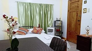 A hotel room with a bed, a desk, a chair, a potted plant, a window with curtains, a wooden door, and a wall-mounted air conditioner.