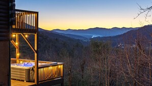 Une terrasse en bois dotée d’un jacuzzi et illuminée de guirlandes lumineuses, offrant une vue sur un paysage montagneux.
