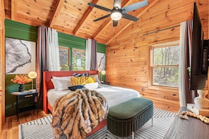 A wooden cabin bedroom with a bed, bedside table, lamp, and a view of the forest through the window.
