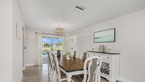 Une salle à manger avec une table en bois, des chaises blanches, un lustre et une vue sur l’extérieur, visible à travers la porte vitrée.
