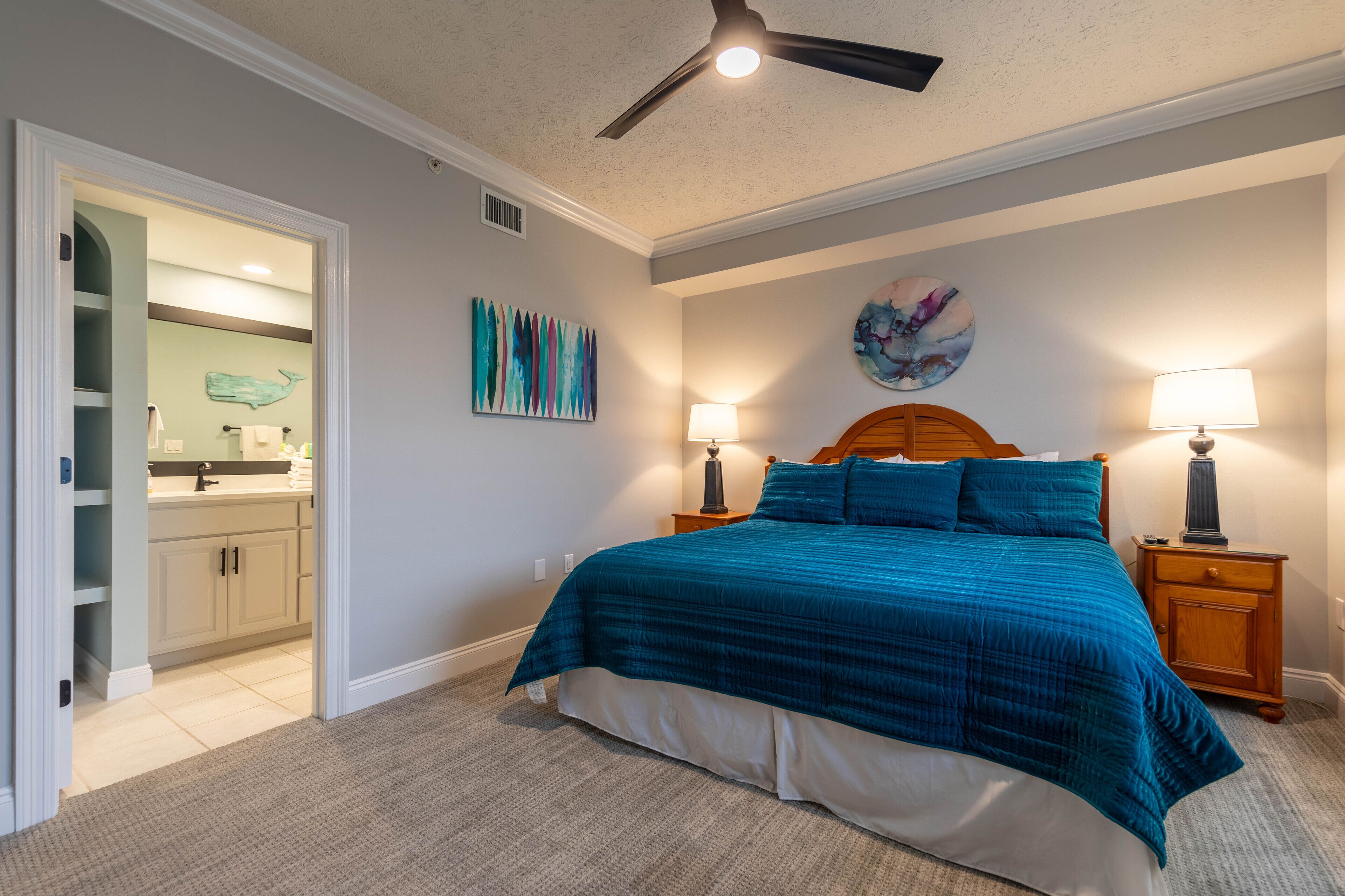 A bedroom with a bed, two bedside tables with lamps, a ceiling fan, and a bathroom visible through an open door.