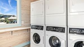A laundry room with stacked washing machines and dryers, a window with a view of a building and palm trees, and wooden paneling on the walls. - 8 Sailfish RV Resort, Port Aransas RV Getaway (Port Aransas)