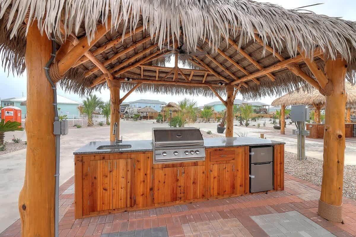 An outdoor kitchen area with a grill, sink, and refrigerator, surrounded by wooden pillars and a thatched roof.