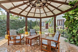 A covered outdoor seating area with a tiled floor, wooden furniture, and a view of a house and greenery.