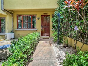 A house with a wooden door, a window with bars, and a small garden with green shrubs.