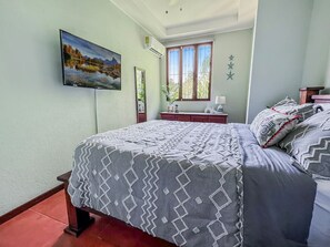 A bedroom with a bed, a TV mounted on the wall, a window with curtains, and a nightstand with a lamp.
