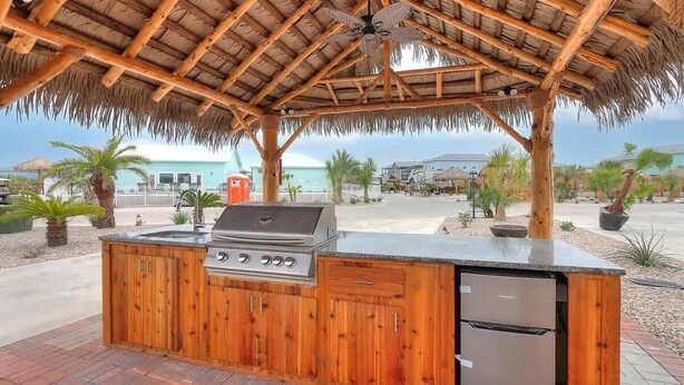 An outdoor kitchen with a grill, refrigerator, and wooden cabinets, under a thatched roof structure. - 34 Sailfish RV Resort, Port Aransas RV Getaway (Port Aransas)
