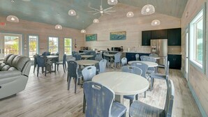 A spacious dining area with round tables and blue chairs, a kitchen area with dark cabinets, and a ceiling fan. - 36 Sailfish RV Resort, Port Aransas RV Getaway (Port Aransas)