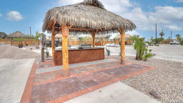 A thatched-roof structure with a wooden bench, surrounded by gravel and a paved area. - 36 Sailfish RV Resort, Port Aransas RV Getaway (Port Aransas)