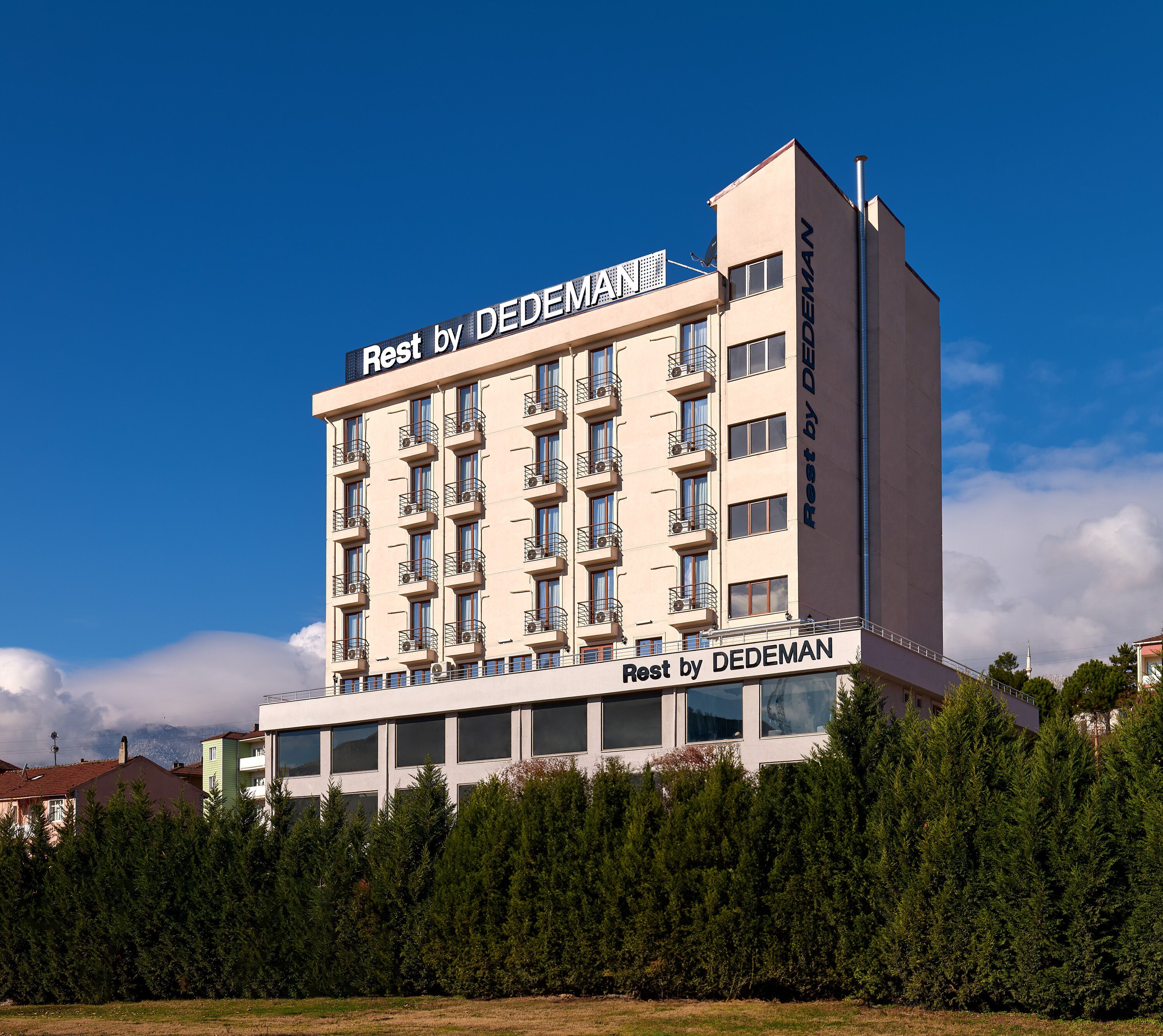 Un hotel de varios pisos llamado 'Rest by DEDEMAN', con balcones y un cielo despejado.