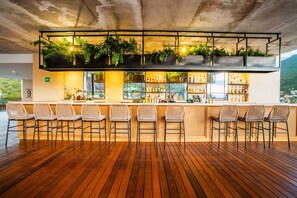 Zona de bar con suelo de madera, taburetes de mimbre y una barra con estantes llenos de botellas.