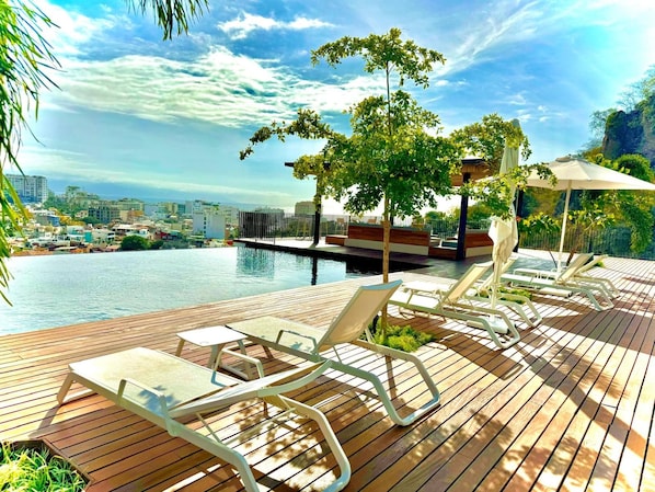 A wooden deck with lounge chairs and a pool, overlooking a cityscape.