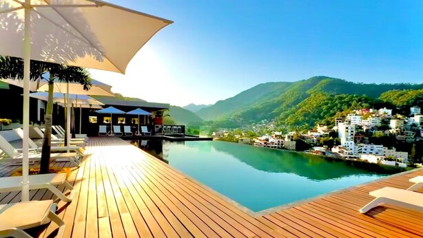 A wooden deck with lounge chairs and umbrellas overlooking a calm body of water and a mountainous landscape.