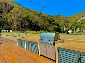 A deck with a barbecue grill and storage cabinets, overlooking a hillside with vegetation and a clear sky.