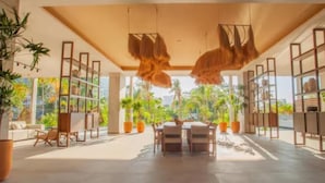 A spacious, sunlit room with a dining area, large windows, and potted plants.