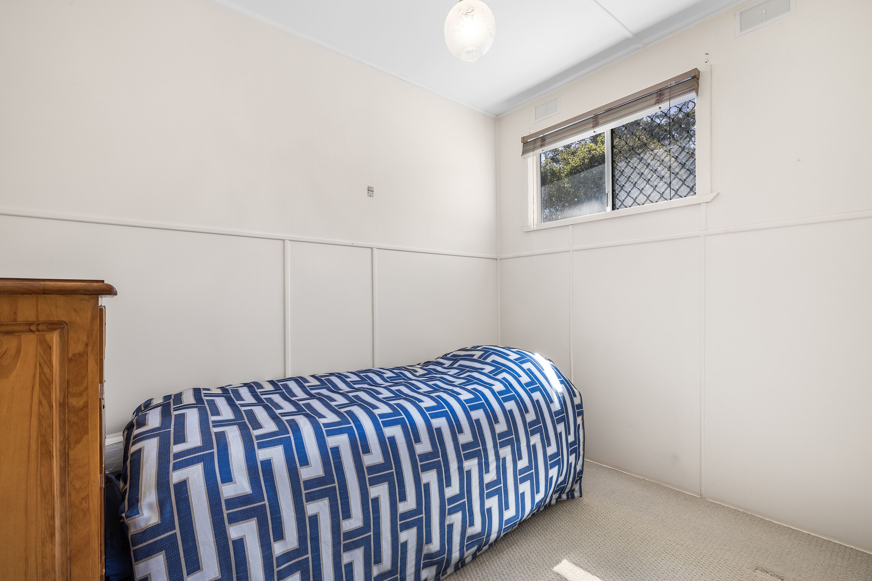A small, single-bed room with a patterned bedspread, a wooden wardrobe, and a window with a view of trees.