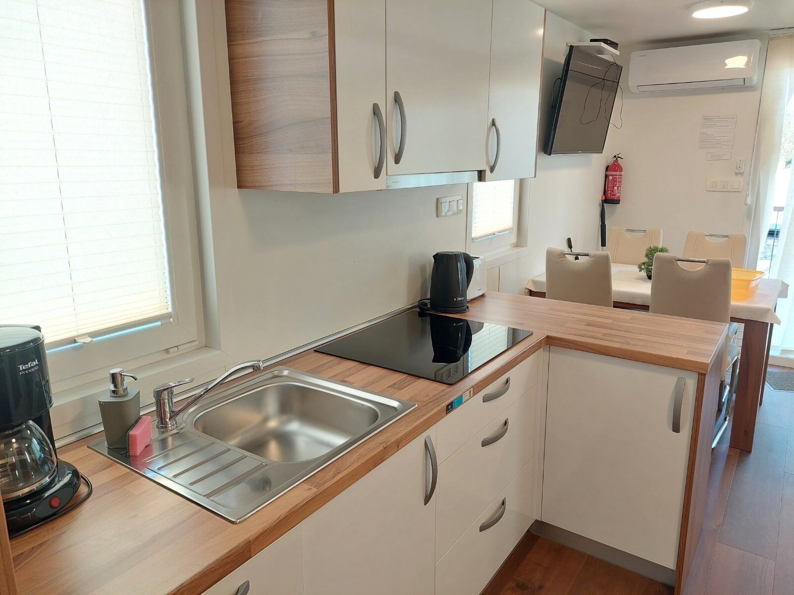 A modern kitchen with white cabinets, a stainless steel sink, a coffee maker, and a flat-screen TV mounted above the counter.