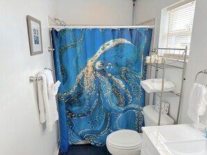 Un baño con cortina de ducha con forma de pulpo azul, un inodoro blanco y un estante metálico con toallas blancas.