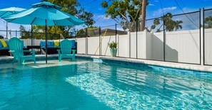 Una piscina con sillas y sombrillas azules, rodeada por una valla blanca y árboles.