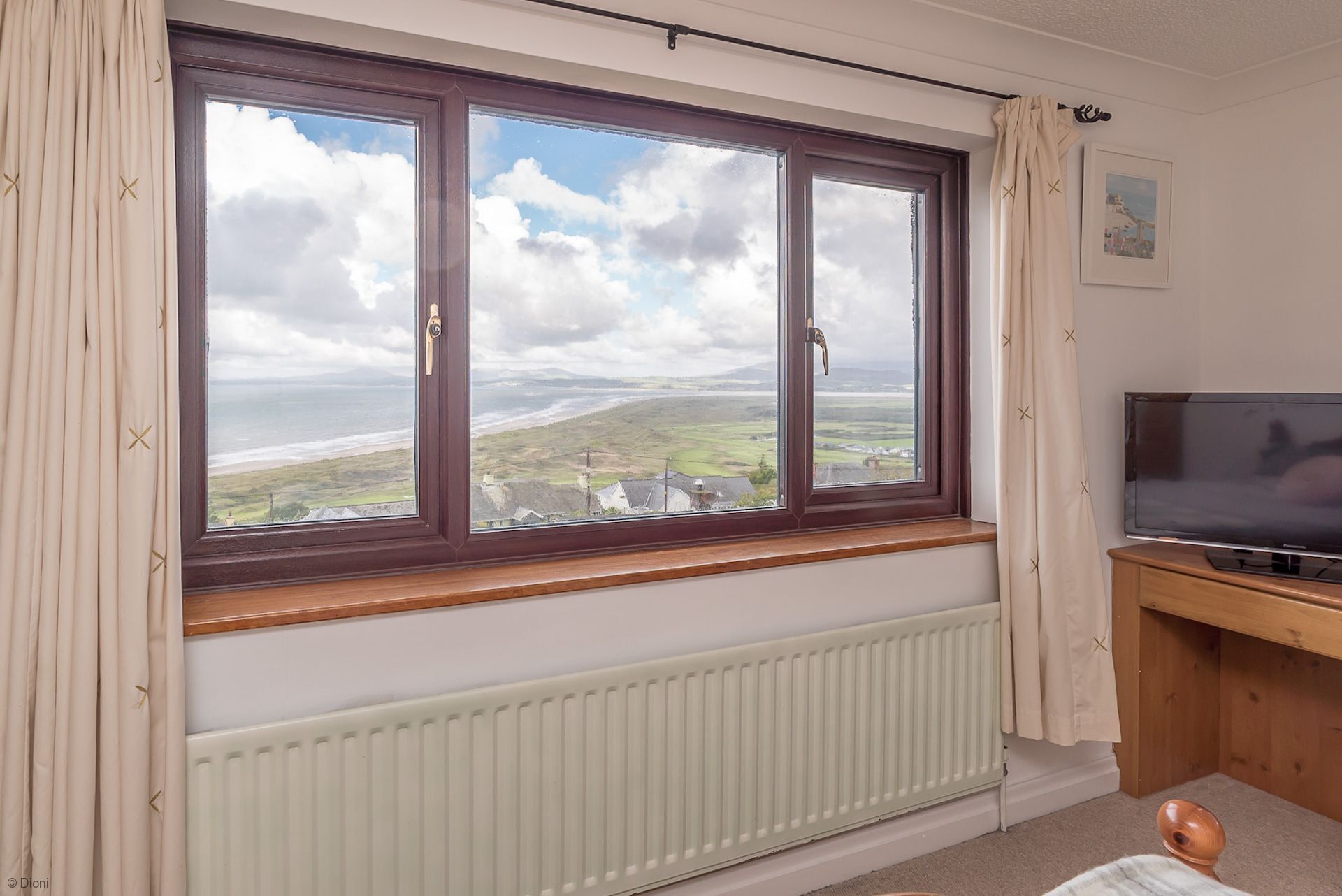 Een kamer met een groot raam dat uitkijkt op een kustlandschap, een radiator beneden en een televisie op een houten standaard.