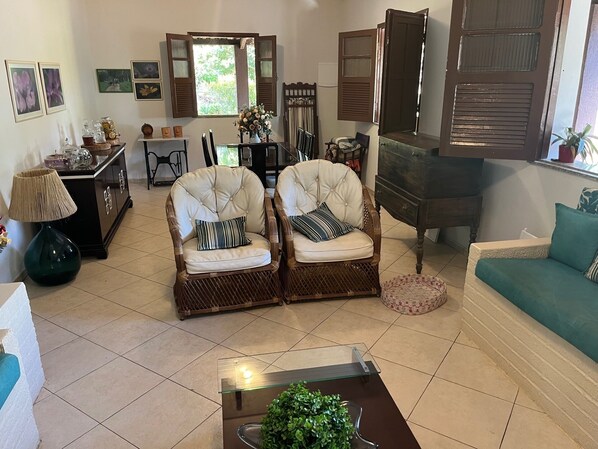 Una sala de estar con dos sillones tapizados, un sofá, una mesa de centro y un comedor con un aparador y una ventana con contraventanas.