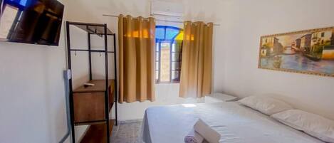 Habitación de hotel con cama, televisión montada en la pared, un cuadro de una escena de canal y una ventana con cortinas.