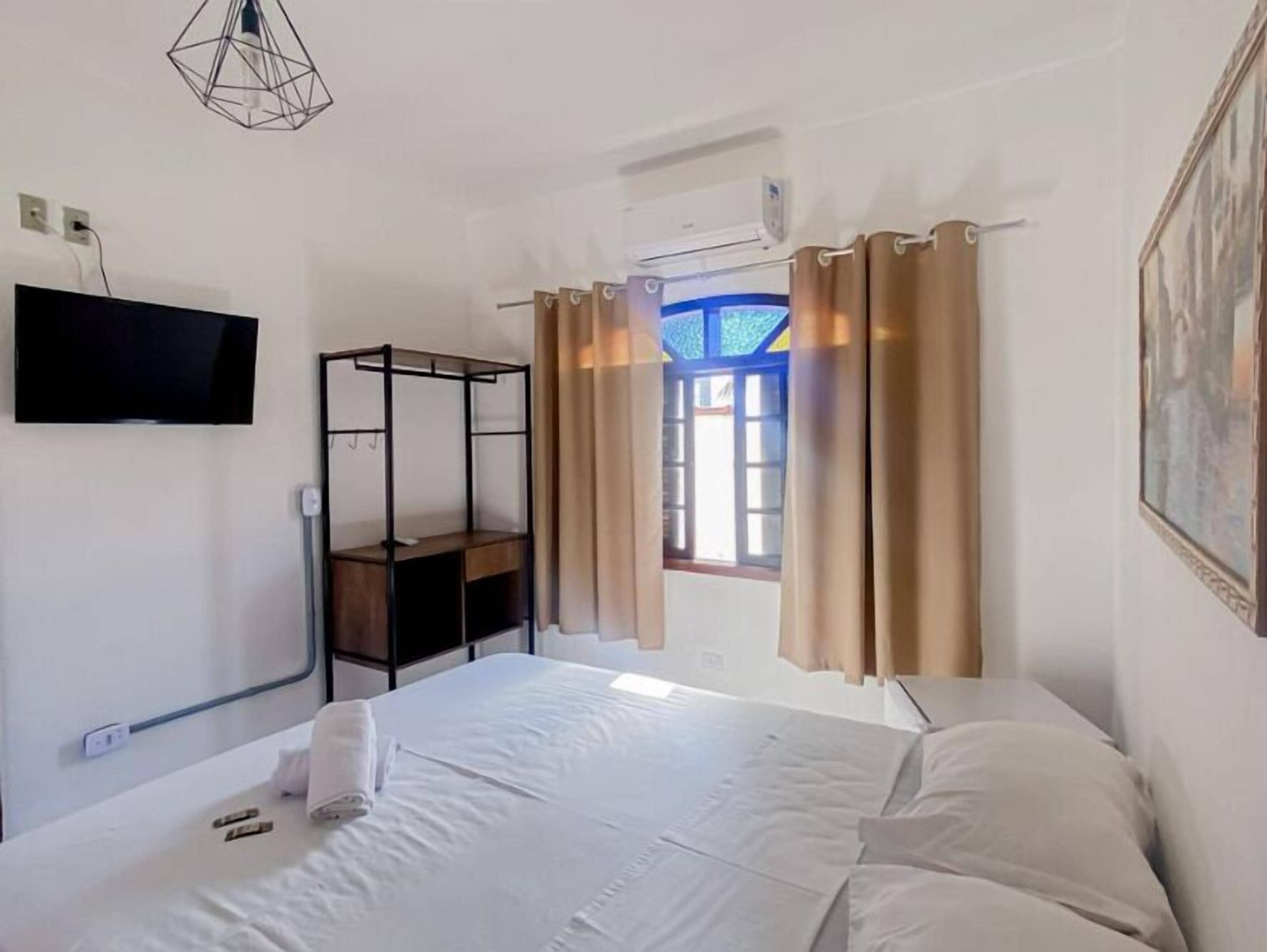 Habitación de hotel con cama, televisor montado en la pared, ventana con cortinas y lámpara colgante.