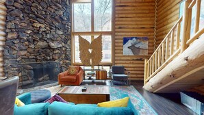 L'intérieur d'un chalet accueillant, avec un mur en pierre, une cheminée, un escalier en bois et une grande fenêtre donnant sur la forêt.