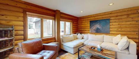 Interior de una cabaña de madera con un sofá de cuero marrón, un sofá seccional blanco y una mesa de centro con libros y controles remotos.