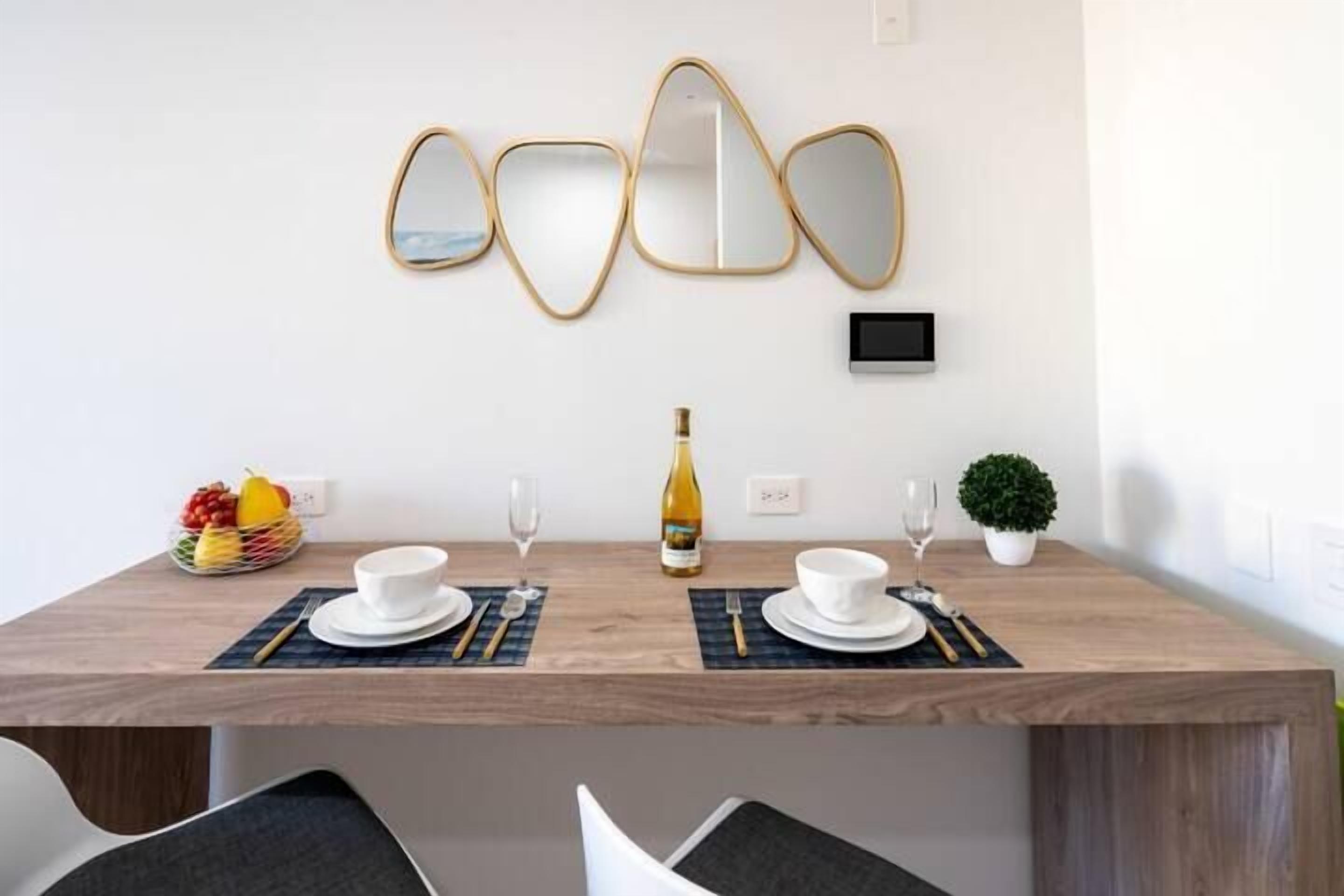 Un comedor con una mesa de madera dispuesta para dos personas, una cesta de frutas, una botella de vino y una pequeña planta en maceta.
