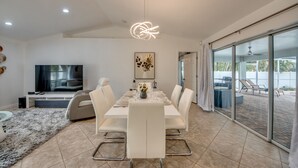 A modern dining area with a large table, white chairs, and a television in the background. - New to VRBO, Pelican Way, Heated Private Pool, Large Lanai area, Billiards..... (Cape Coral)