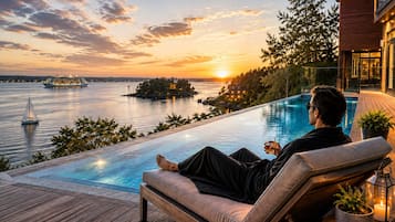 A person relaxing on a deck chair by an infinity pool overlooking a body of water with a sunset in the background.