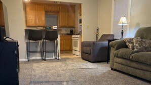 A living room with a grey carpet, a sofa, a coffee table, and a TV stand. There is a kitchen area with wooden cabinets and a stove. - Private stand-alone little house.   (Fairview Park)