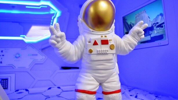 A figure in a white astronaut suit with a gold helmet, standing in a room with blue lighting and a control panel in the background. - Cosmos capsule coworking hub London (London)