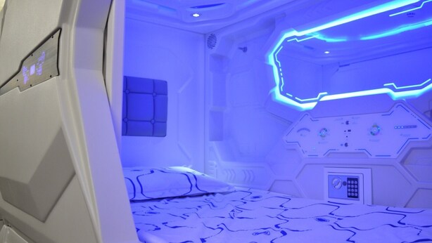 Ein futuristisches Zimmer mit einem Bett, Bedienfeldern und einer blau beleuchteten Decke.
