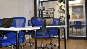A room with blue chairs, a white table, and a laptop. There is a shelf with bottles and a plant in the background. - Cosmos capsule coworking hub London (London)