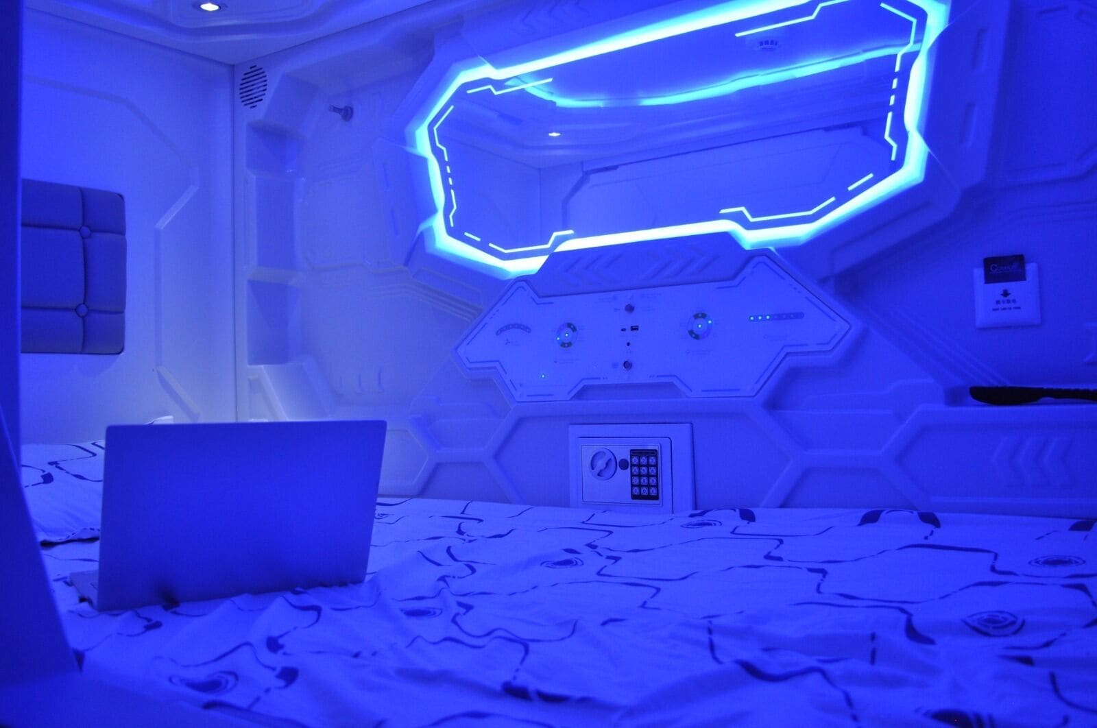 A room with a bed, a laptop on the bed, and a wall with a geometric light fixture.