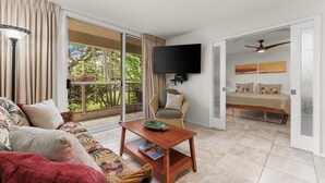 A room with a sofa, a chair, a table, a TV, and a bed with a view of greenery. - Luxury Condo at Kameole Beach (Kihei)