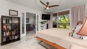 A bedroom with a bed, a bookshelf, a ceiling fan, a TV, and a view of the outdoors. - Luxury Condo at Kameole Beach (Kihei)