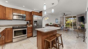 A modern kitchen with wooden cabinets, stainless steel appliances, a large island with stools, and a dining area with a view. - Luxury Condo at Kameole Beach (Kihei)