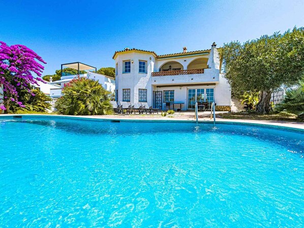 A poolside view of a two-story house with a clear blue pool, surrounded by lush greenery and flowering plants.