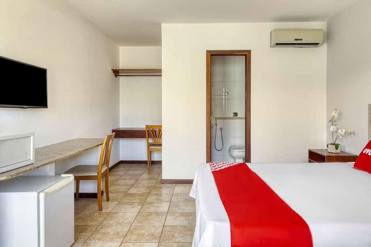 Une chambre d’hôtel équipée d’un lit, d’un bureau, d’une chaise, d’une télévision, d’un réfrigérateur, d’une douche et d’une petite table sur laquelle est posé un vase de fleurs.