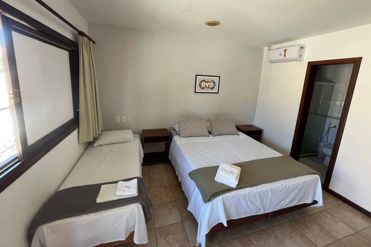 A hotel room with two single beds, a large window with curtains, a wall-mounted air conditioner, and a framed picture.